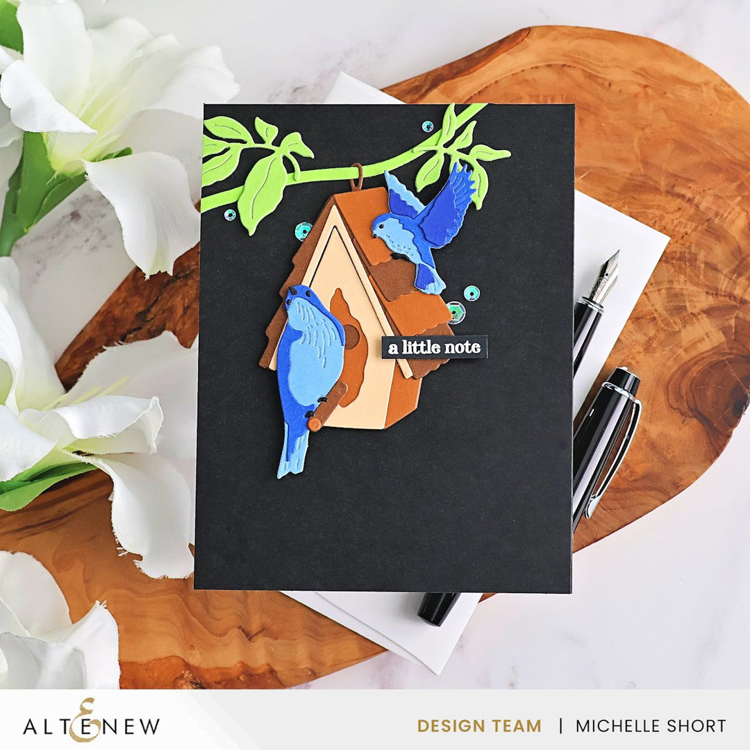 Black card with blue bird and wooden house design on a wooden surface with flowers and stationery.