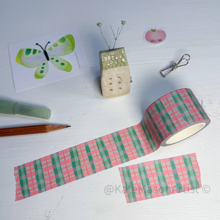 Pink and green plaid washi tape shown on a white wooden surface, with a roll of the tape, a cut strip, and small styled craft props including a butterfly card, pin cushion, clip, pencil, and marker.