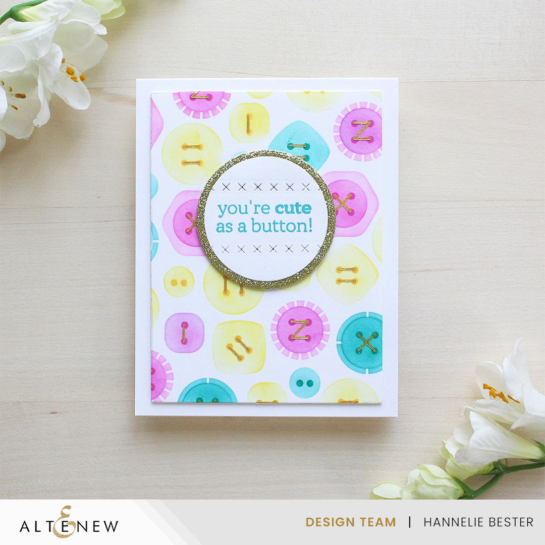 Card with colorful button pattern and 'you're cute as a button!' text on a light wooden surface with flowers.