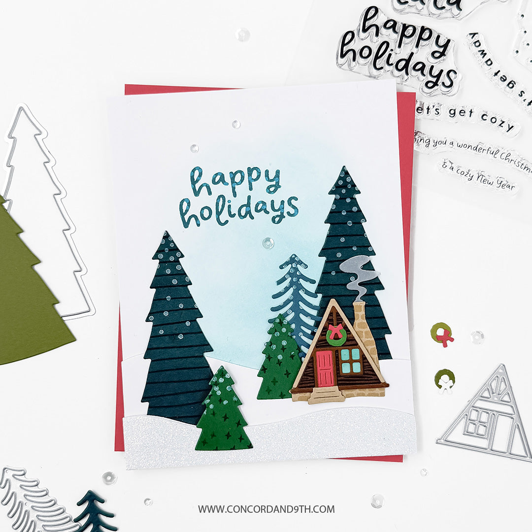 Concord & 9th Into the Woods Dies — the cabin and layered pine tree scene in a snowy landscape, with a "happy holidays" sentiment.