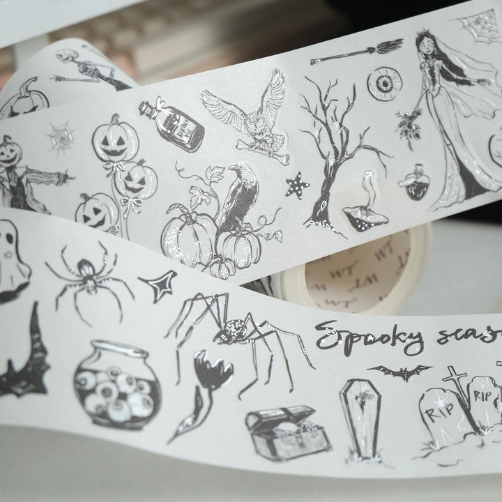 A roll of white washi tape with black spooky illustrations, including pumpkins, a witch, a spider, a potion bottle, bats, and tombstones, shown partially unrolled.