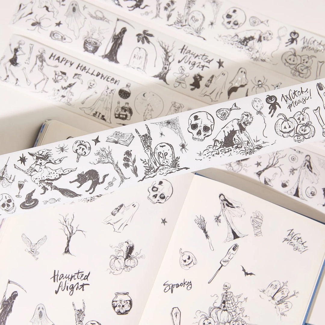 Halloween-themed washi tape rolls with black line-art illustrations of skulls, ghosts, pumpkins, bats, and spooky text, shown partially unrolled over open notebooks.