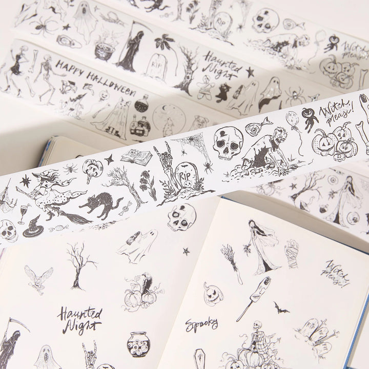 Halloween-themed washi tape rolls with black line-art illustrations of skulls, ghosts, pumpkins, bats, and spooky text, shown partially unrolled over open notebooks.