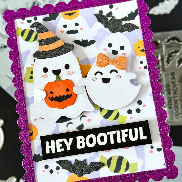 A handmade Halloween greeting card with a glittery purple scalloped border, featuring cute ghost cutouts, bats, candy, and a jack-o'-lantern. The card has a black banner at the bottom with the text 'HEY BOOTIFUL' in white.