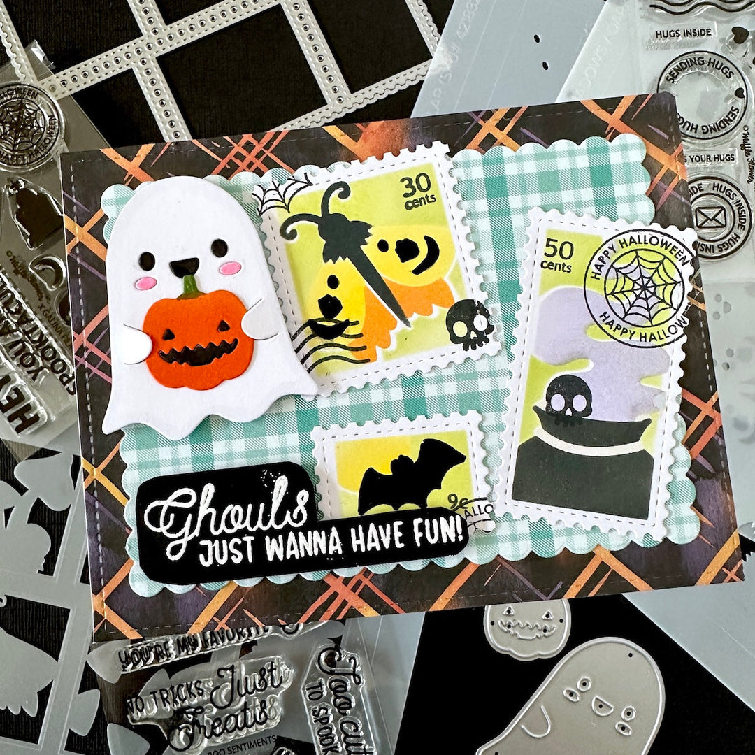A Halloween-themed craft card displayed on a black background with craft stencils and clear stamps around it. The main product features a ghost holding a jack-o'-lantern, postage-style stamps with pumpkins, bats, skulls, spiderwebs, and the phrase 'Ghouls JUST WANNA HAVE FUN!'.