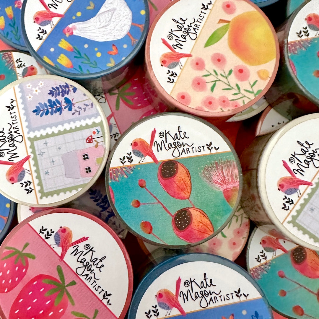 A close-up, top-down view of Washi tapes with illustrated labels reading 'Kate Mason Artist.' The labels feature colourful floral and bird motifs, including strawberries, peaches, blue birds, pink flowers, and pastel landscape panels.