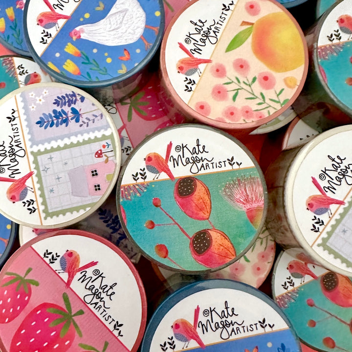 A close-up, top-down view of Washi tapes with illustrated labels reading 'Kate Mason Artist.' The labels feature colourful floral and bird motifs, including strawberries, peaches, blue birds, pink flowers, and pastel landscape panels.