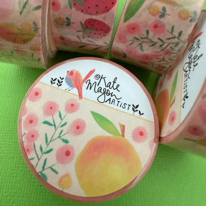 A round roll of decorative washi tape with a fruit and floral design, shown in front of other rolls of the same pattern on a bright green surface. The label reads 'Kate Mason Artist'.