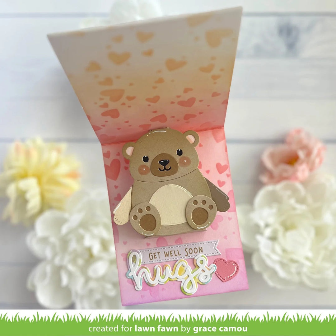 A handmade pop-up greeting card featuring a cute brown teddy bear with a cream belly, set against a pink background with heart patterns and the words 'GET WELL SOON hugs'. The card is displayed open with decorative flowers in the background.