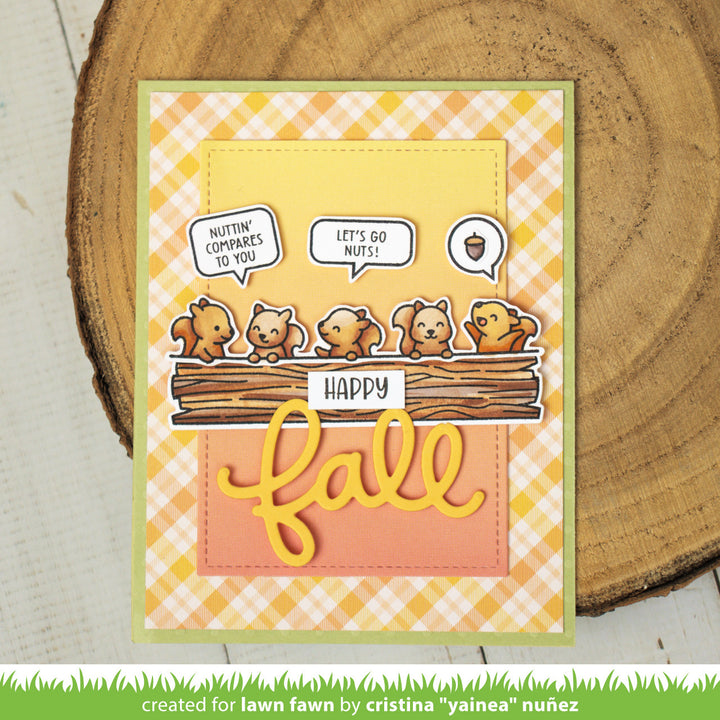Autumn-themed card with cartoon squirrels and 'Happy Fall' text on a checkered background, placed on a wooden surface.