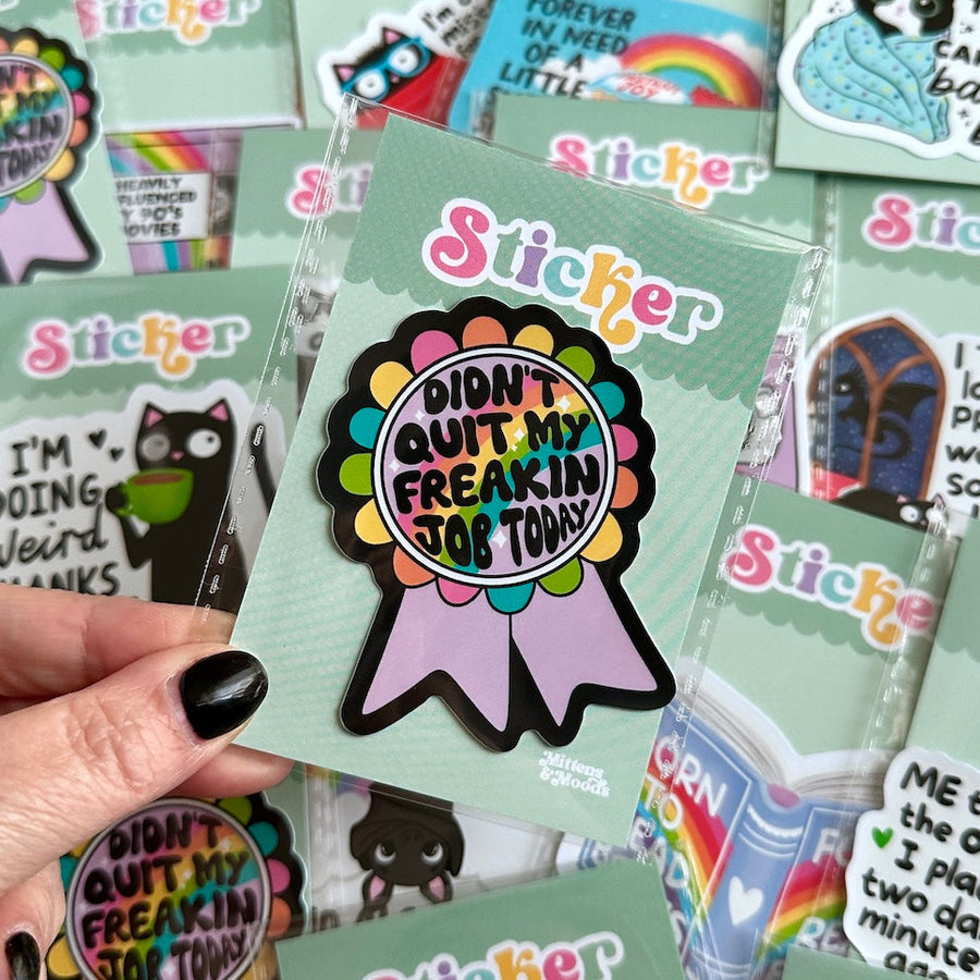 A hand with black nail polish holds a round sticker on a mint green backing card. The sticker has a colorful scalloped circle with the text 'I DIDN'T QUIT MY FREAKING JOB TODAY!' in the center, attached to a lavender ribbon.