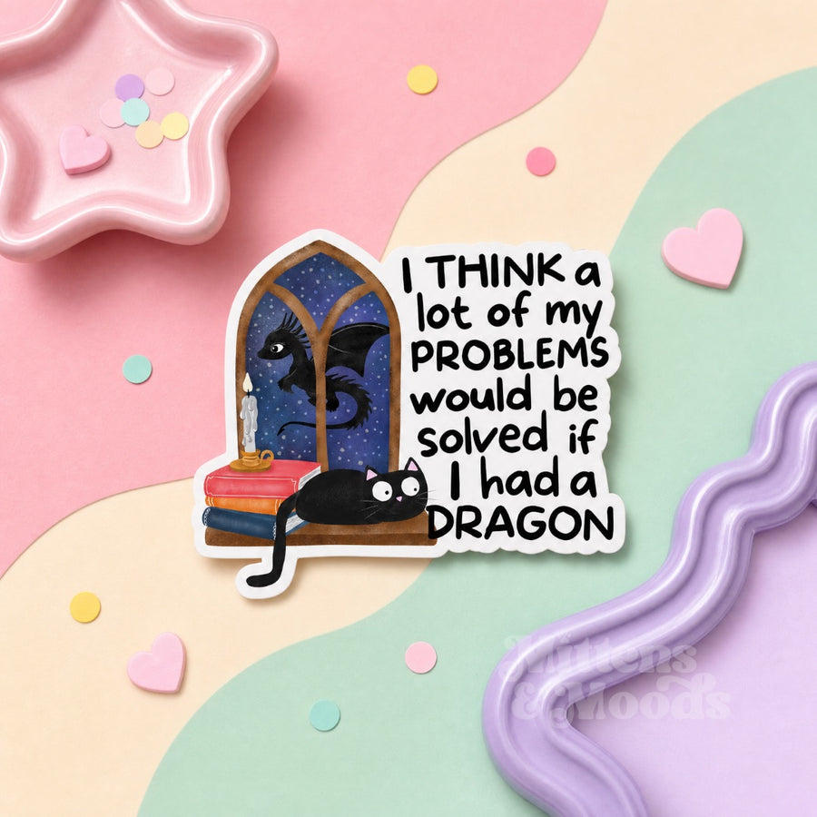 A die-cut sticker featuring a black cat with a starry night sky window and a black dragon, next to the text 'I THINK a lot of my PROBLEMS would be solved if I had a DRAGON'. The sticker is displayed on a pastel wavy background with a pink star-shaped dish and pastel confetti.