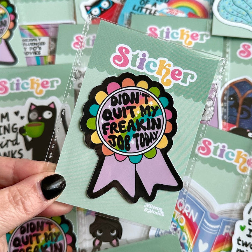 A hand with black nail polish holds a round sticker on a mint green backing card. The sticker has a colorful scalloped circle with the text 'I DIDN'T QUIT MY FREAKING JOB TODAY!' in the center, attached to a lavender ribbon.
