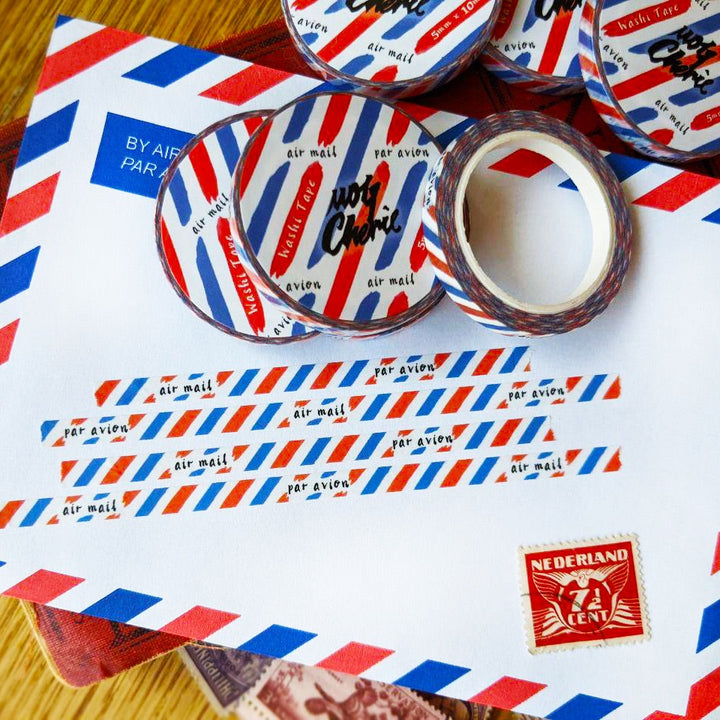 A set of decorative washi tapes with a red, white, and blue retro air mail stripe pattern, shown on and around a vintage-style white airmail envelope on a wooden surface.