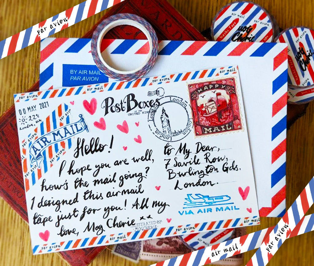A styled flat-lay of vintage-themed postal stationery on a wooden surface, featuring a white postcard with red and blue striped borders, handwritten lettering, pink hearts, a red vintage stamp, and a blue 'AIR MAIL' emblem. A roll of matching patterned tape and additional red, white, and blue striped tape are arranged around it.