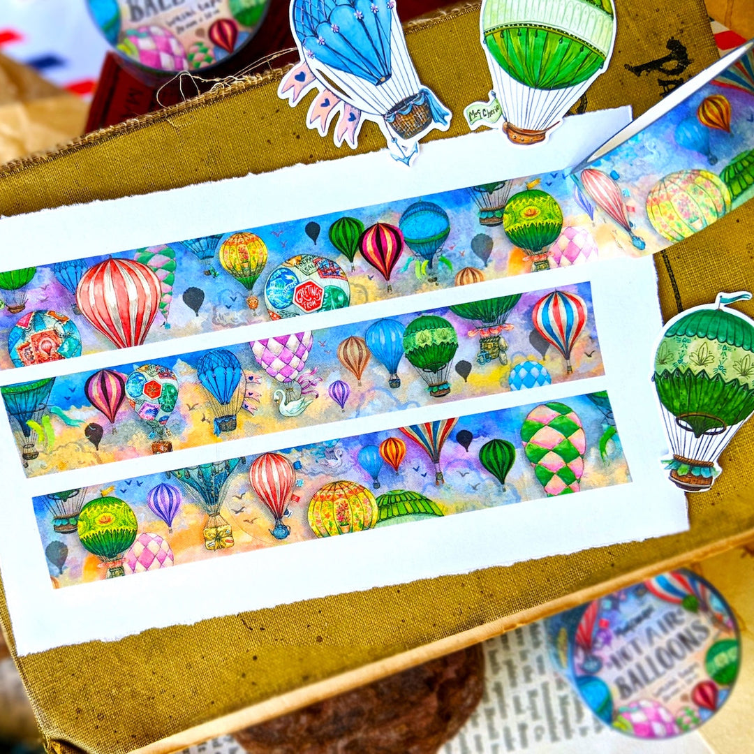 Three horizontal strips of washi tape featuring a colorful watercolor illustration of hot air balloons, displayed on a kraft paper box with other decorative items.