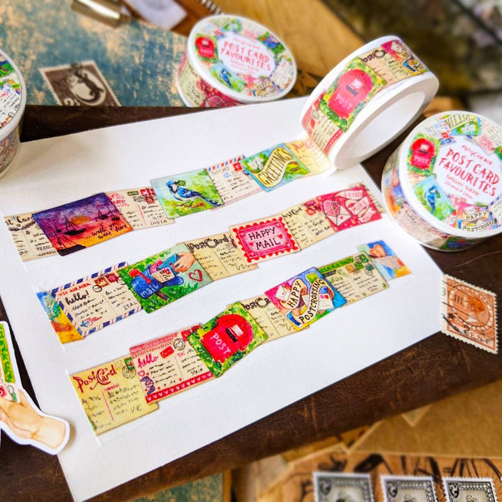 A styled flat-lay of decorative washi tape rolls and strips featuring vintage postcard designs, shown on a white sheet over a dark wooden surface with postage stamps and gift tags nearby.