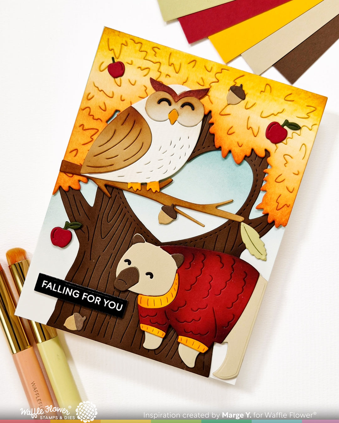 A handmade layered card featuring an owl and bear in autumn foliage, styled on a white surface with craft pens and paper swatches. The card reads 'FALLING FOR YOU' and is inspired by Marge Y.