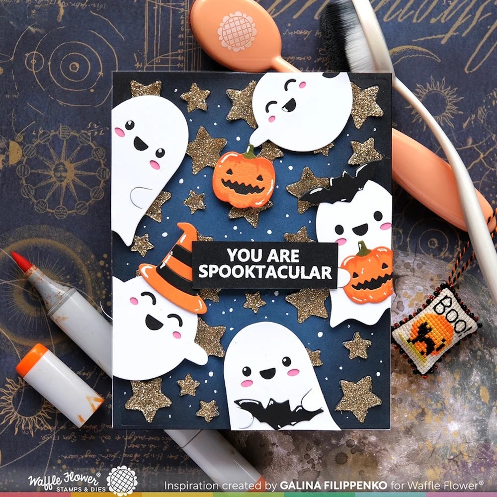 A Halloween-themed handmade greeting card with cute cartoon ghosts, jack-o'-lanterns, glittery stars, and the text 'YOU ARE SPOOKTACULAR' on a dark blue background. The card is styled in a flat-lay with craft tools and markers on a dark patterned surface.