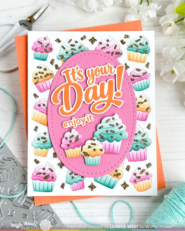 A handmade greeting card featuring a pink oval with the words 'It's your Day! enjoy it' in orange and white script, surrounded by colorful illustrated cupcakes. The card is displayed on a white wooden surface with crafting tools and flowers in the background.
