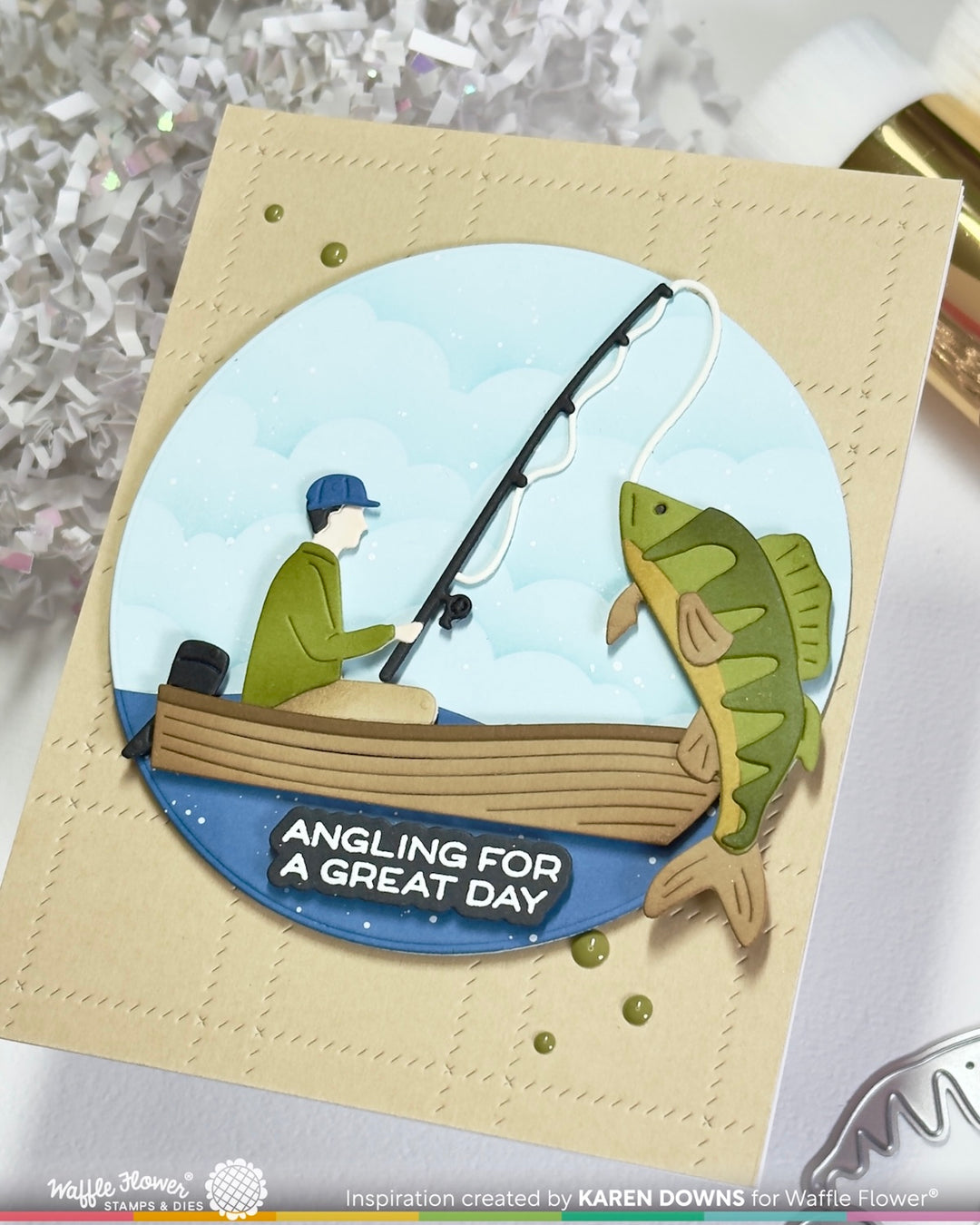A handmade greeting card featuring a die-cut illustration of a man fishing with a large fish on a boat, with the text 'ANGLING FOR A GREAT DAY'. The card is made from beige cardstock with a circular blue and green design layered on top, photographed on a white surface with decorative tinsel in the background.