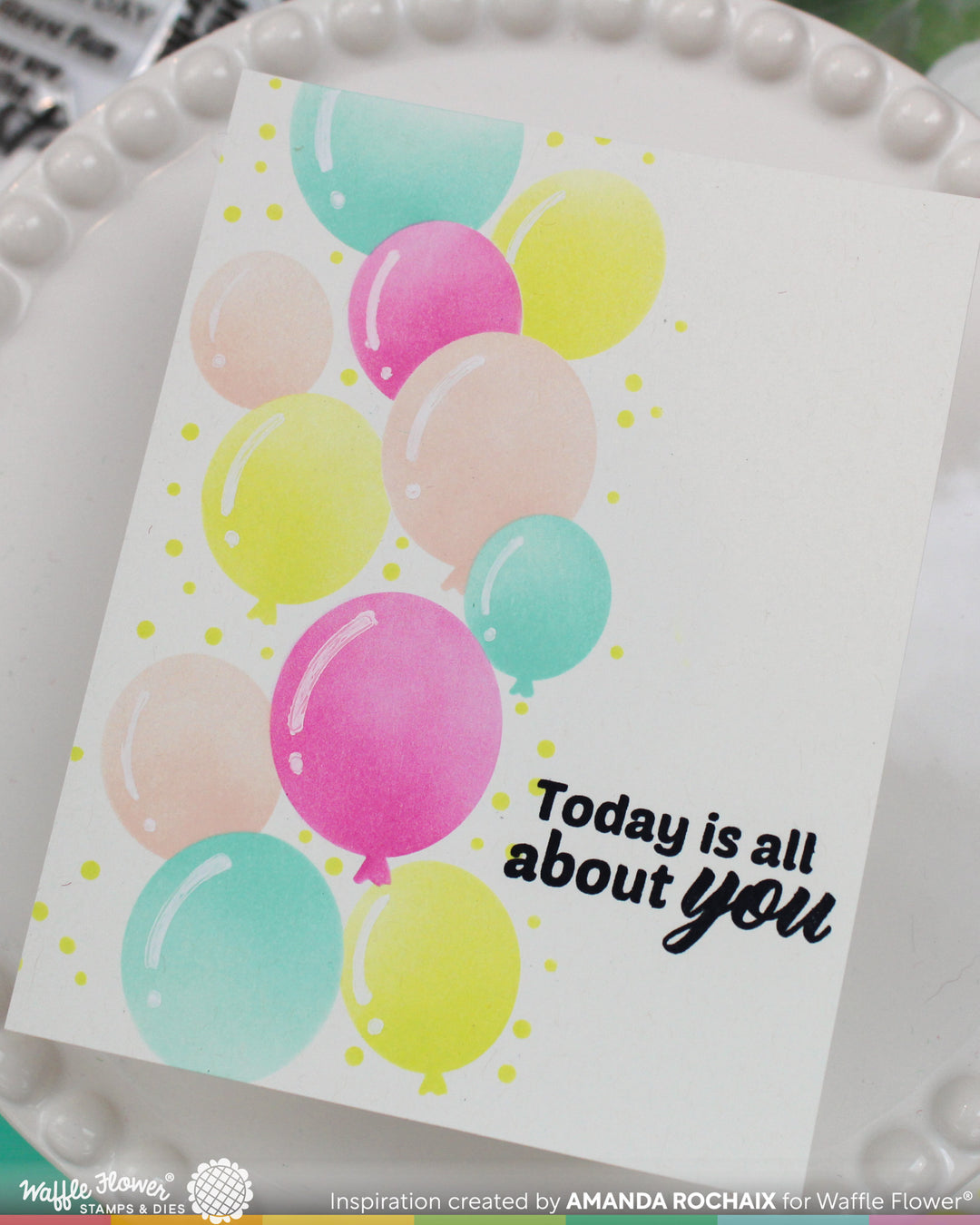 A handmade greeting card featuring watercolor balloons in pastel pink, yellow, and teal on a white background, with the black script text 'Today is all about you'. The card is displayed on a white beaded dish.