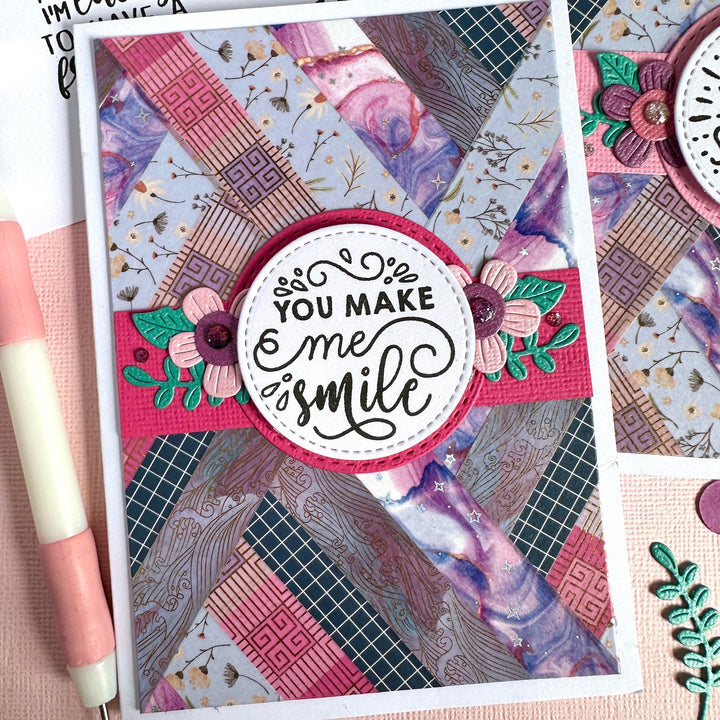 A handmade layered card featuring a washi tape patchwork collage of pink, purple, and blue with a central white circular badge that reads 'YOU MAKE me smile' in black script. The card is styled on a pink surface next to a pink and white pen.
