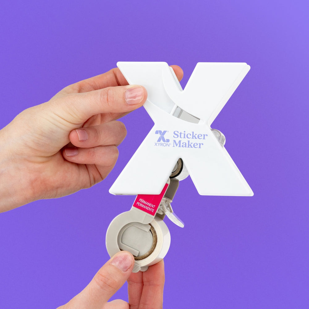 Hand holding a Xyron X-Sticker Maker device against a purple background