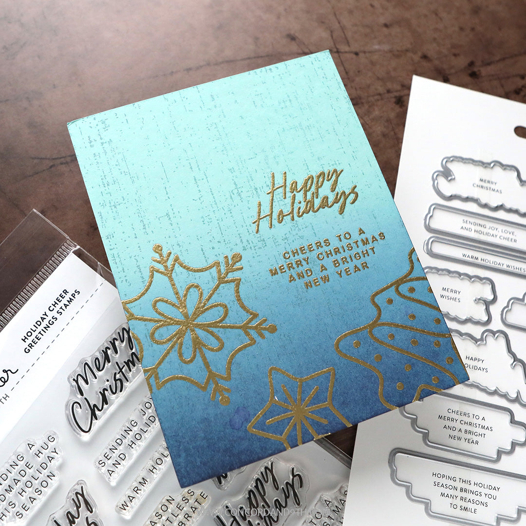 Concord & 9th Kristina Werner Holiday Cheer Greetings Stamps — a teal-to-blue ombre "Happy Holidays" card with gold-embossed snowflakes and a layered sentiment.
