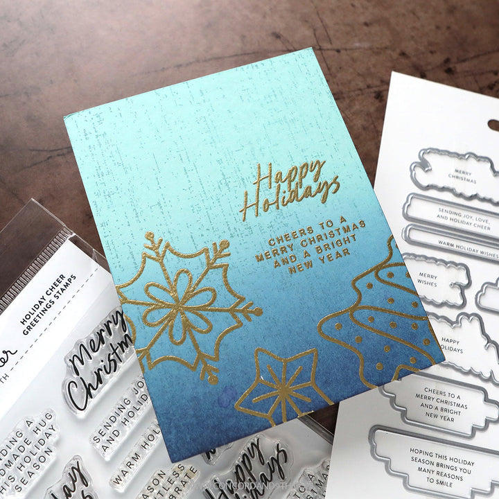 Concord & 9th Kristina Werner Holiday Cheer Greetings Stamps — a teal-to-blue ombre "Happy Holidays" card with gold-embossed snowflakes and a layered sentiment.