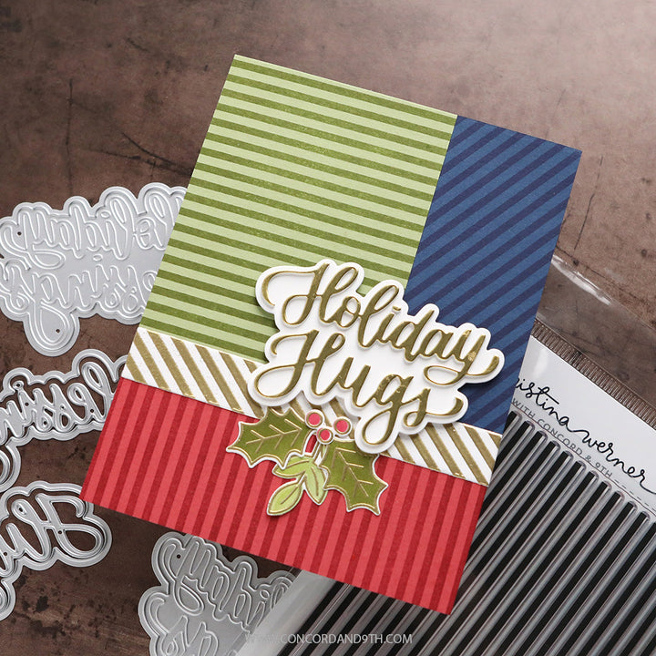 Concord & 9th Kristina Werner XL Basic Stripes Background Stamp — a multicolor striped "Holiday Hugs" card with a stamped holly sprig accent.