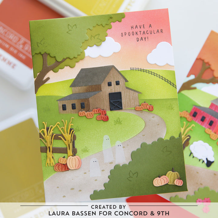 A layered paper-cut style greeting card featuring an autumn farm scene with a barn, pumpkins, ghosts, trees, and the text 'HAVE A SPOOKTACULAR DAY!'. The card is photographed on a light surface with other cards partially visible around it.