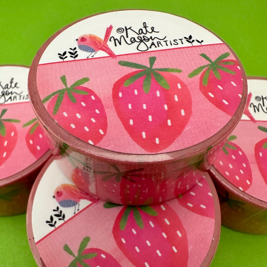 A round container of washi tape featuring a pink strawberry pattern, with a white top label that reads 'Kate Mason Artist' and shows a small bird illustration, set against a bright green background.