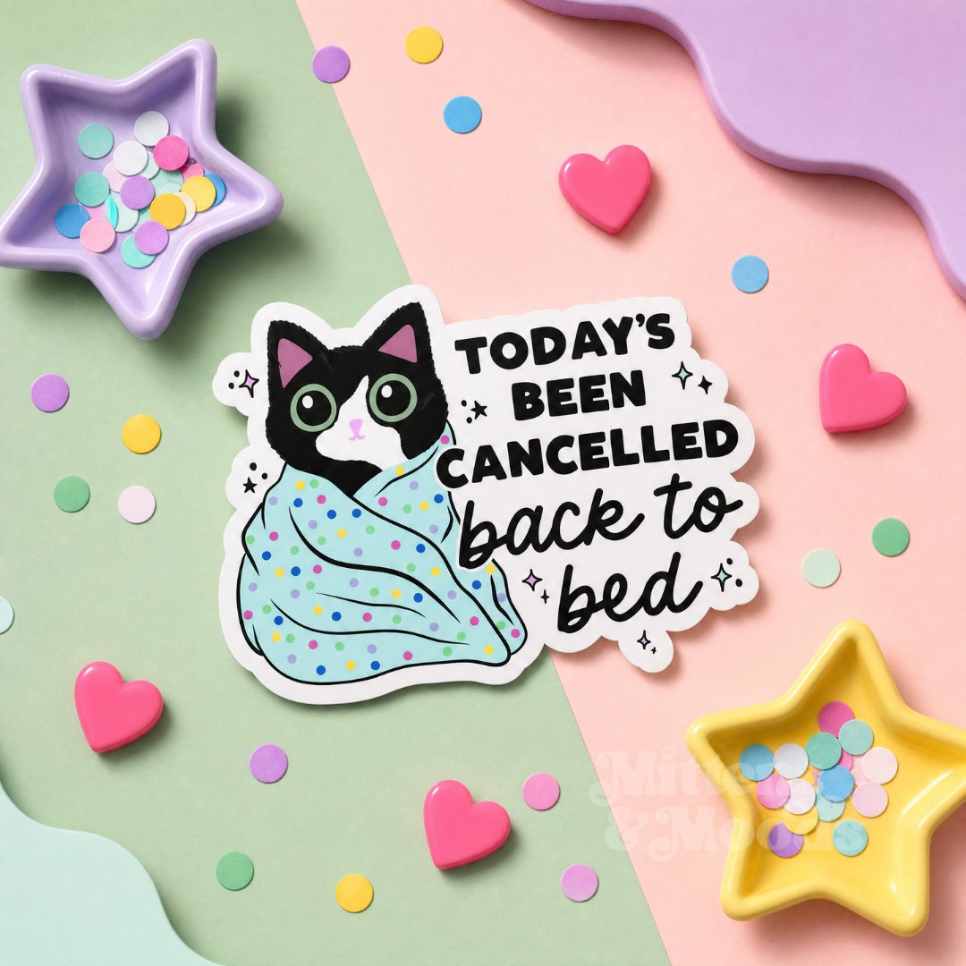 A die-cut sticker featuring a black-and-white cat wrapped in a pastel polka-dot blanket with the text 'TODAY'S BEEN CANCELLED back to bed,' styled on a pastel background with star-shaped confetti dishes and heart-shaped props.
