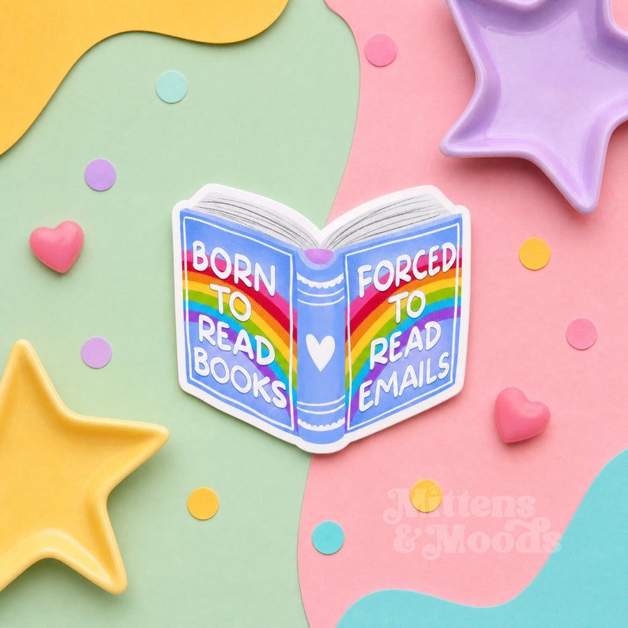 A die-cut sticker shaped like an open book with a rainbow on the cover and the text 'BORN TO READ BOOKS FORCED TO READ EMAILS' in white lettering, shown on a pastel flat-lay background with star and heart props. The brand name 'Mittens & Moods' appears in the lower right.