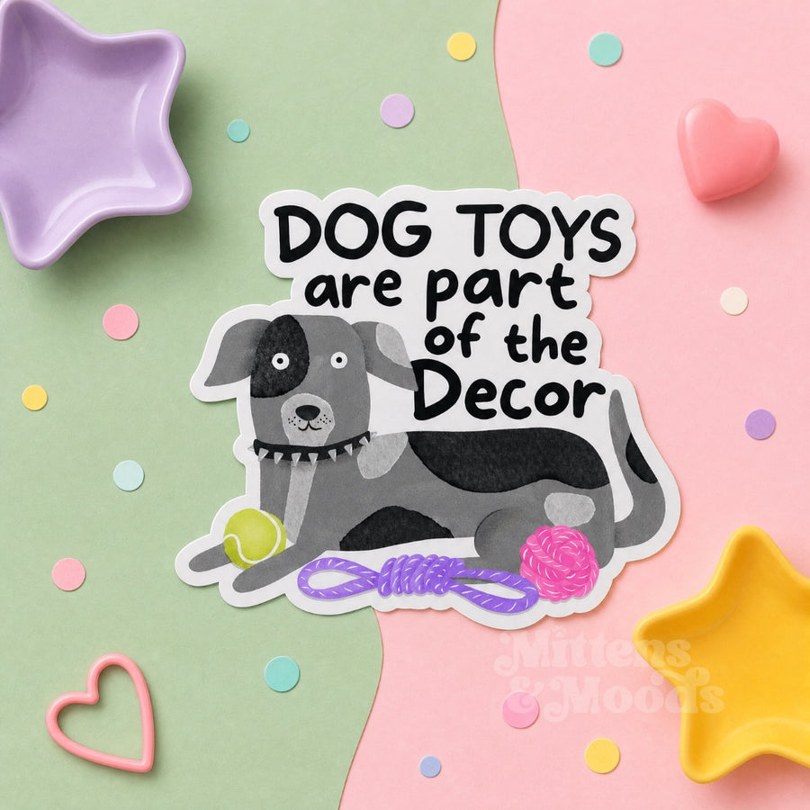 A die-cut sticker featuring a cartoon gray and black dog lying on a pink and mint green background with pastel confetti and decorative props. The sticker reads “DOG TOYS are part of the Decor” in bold black text.