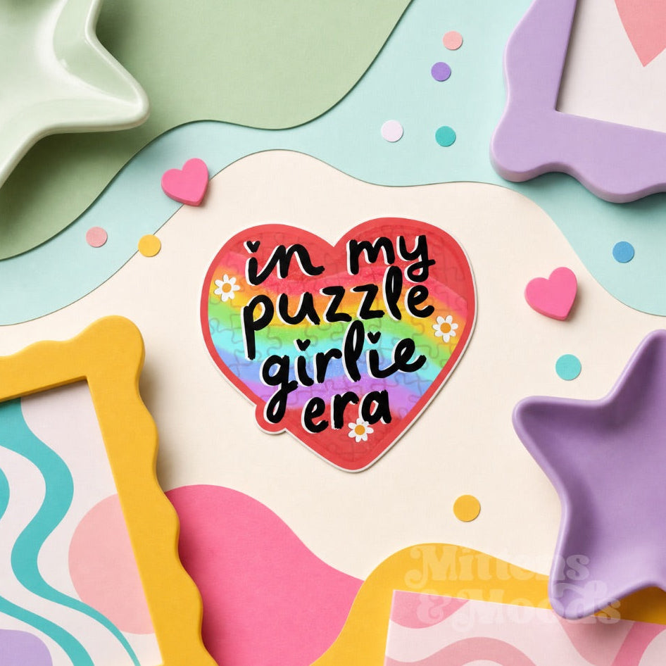A heart-shaped sticker with the text 'in my puzzle girlie era' in black script over a rainbow gradient background, surrounded by pastel decorative paper cutouts, star and frame shapes, and small confetti dots.