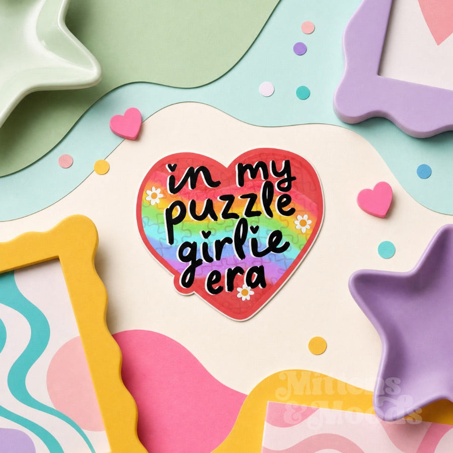 A heart-shaped sticker with the text 'in my puzzle girlie era' in black script over a rainbow gradient background, surrounded by pastel decorative paper cutouts, star and frame shapes, and small confetti dots.