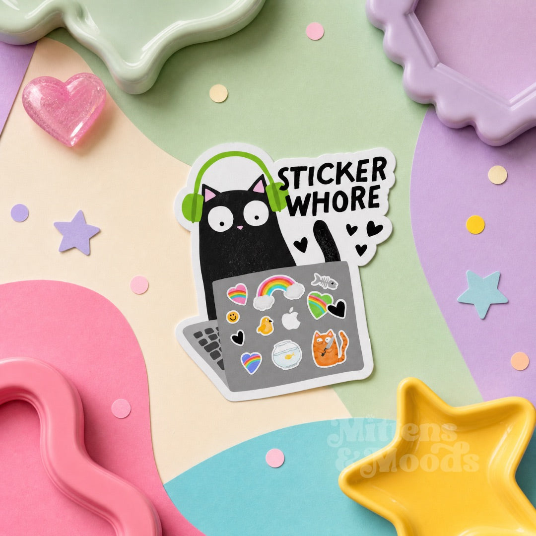A die-cut sticker featuring a black cat wearing green headphones and looking at a laptop covered in colorful stickers, with the text 'STICKER WHORE' in white. The sticker is displayed on a pastel-colored flat-lay background with star and heart-shaped props.