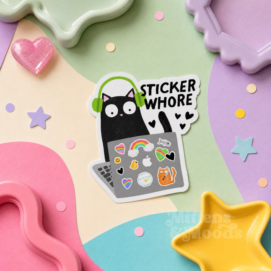 A die-cut sticker featuring a black cat wearing green headphones and looking at a laptop covered in colorful stickers, with the text 'STICKER WHORE' in white. The sticker is displayed on a pastel-colored flat-lay background with star and heart-shaped props.